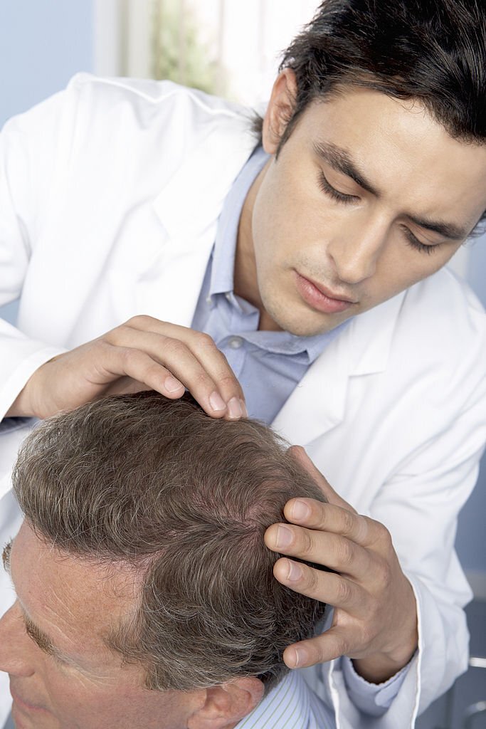 Hair transplant consultation. Trichologist examining the scalp of a man who is contemplating hair transplant surgery. The doctor is checking for areas of male pattern baldness (alopecia).